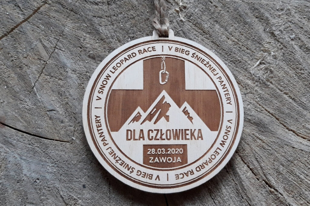 Okrągły drewniany medal z grawerem  ostrych górskich szczytów, napisem V Bieg Śnieżnej Pantery oraz logotypem wydarzenia.