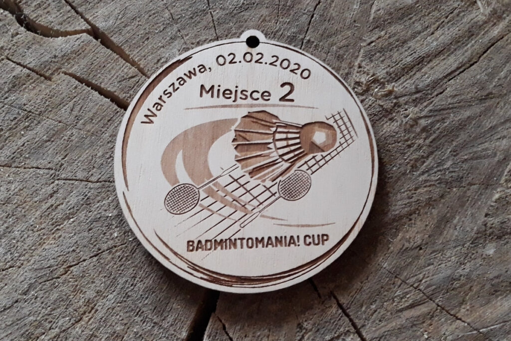 Okrągły drewniany medal na turniej badmintona, z precyzyjnym grawerem lotki oraz dwóch rakiet i siatki, wykonany z jasnej brzozy o krystalicznie czystej powierzchni bez osmaleń. U góry zajęte miejsce oraz miejscowość i data.