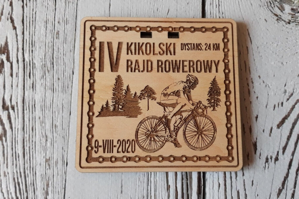 Kwadratowy drewniany medal na „Kikolski Rajd Rowerowy”, z dynamicznym grawerem rowerzysty na trasie, nazwą wydarzenia oraz motywami roślinnymi, wykonany z jasnej brzozy bez śladów dymu i osmaleń. Dookoła ramka z łańcucha rowerowego.