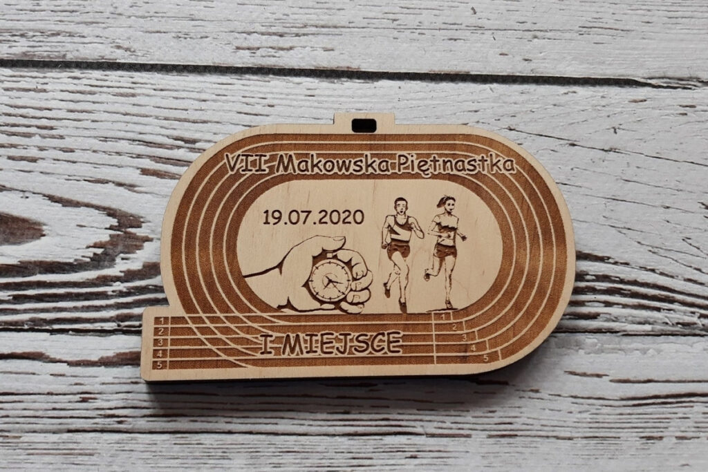 Drewniany medal na bieg „Makowska Piętnastka”, z grawerem imitującym tory bieżni lekkoatletycznej, sylwetką biegaczy oraz ręką trzymającą stoper - mierzącą czas, wykonany z czystej brzozy bez osmaleń.