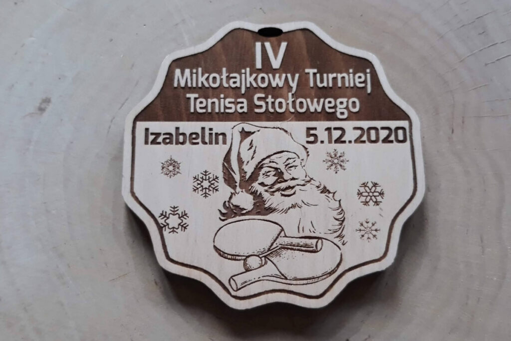 Okrągły drewniany medal na Mikołajkowy Turniej Tenisa Stołowego, z grawerem rakietki, piłeczki oraz twarzy i czapki św. Mikołaja, wykonany z płyty - jasnej brzozy bez śladów dymu.