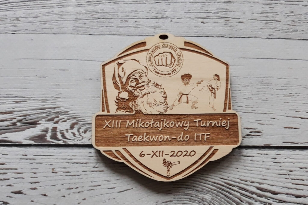 Drewniany medal w kształcie tarczy na „Mikołajkowy Turniej Taekwon-do”, z grawerem zawodnika , św. Mikołaja w czapce oraz logiem federacji, wykonany z czystej brzozy bez śladów osmaleń.