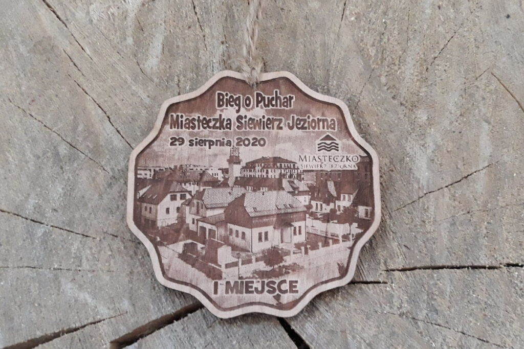 Okrągły drewniany medal z precyzyjnie wygrawerowaną panoramą miasteczka Siewierz Jeziorna, przedstawiający charakterystyczną architekturę domów nad wodą, wykonany z jasnej brzozy bez śladów dymu i osmaleń.