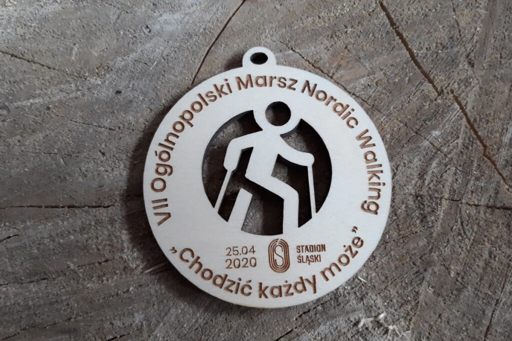 Okrągły drewniany medal na Ogólnopolski Rajd Nordic Walking „Chodzić każdy może”, z ażurowo wyciętą sylwetką osoby maszerującej z kijkami oraz grawerem logotypu wydarzenia, wykonany z czystej brzozy bez śladów dymu.