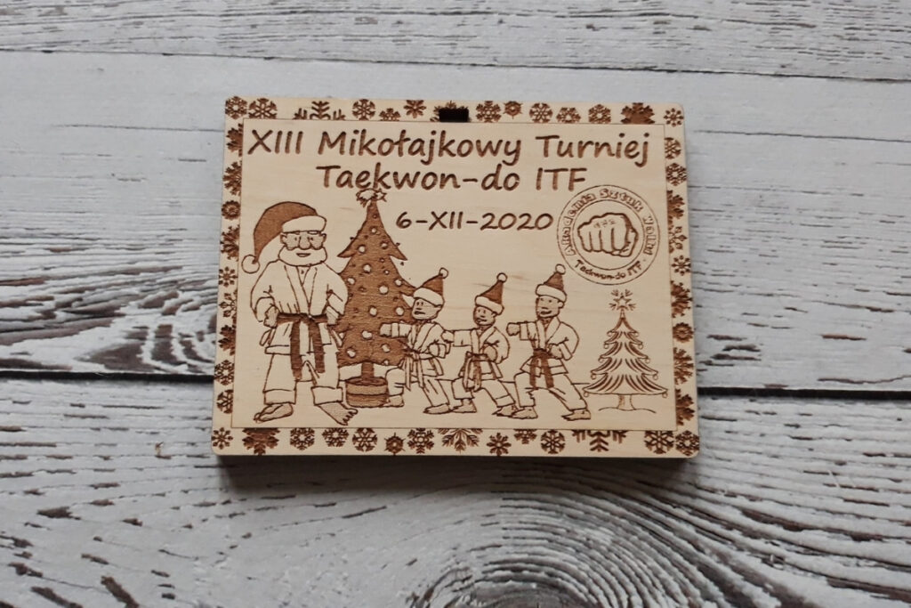Prostokątny drewniany medal na XIII Mikołajkowy Turniej Taekwon-do ITF z grawerem zawodnika wykonującego kopnięcie boczne, czapką św. Mikołaja i logotypem federacji, czyste wykończenie bez osmaleń.