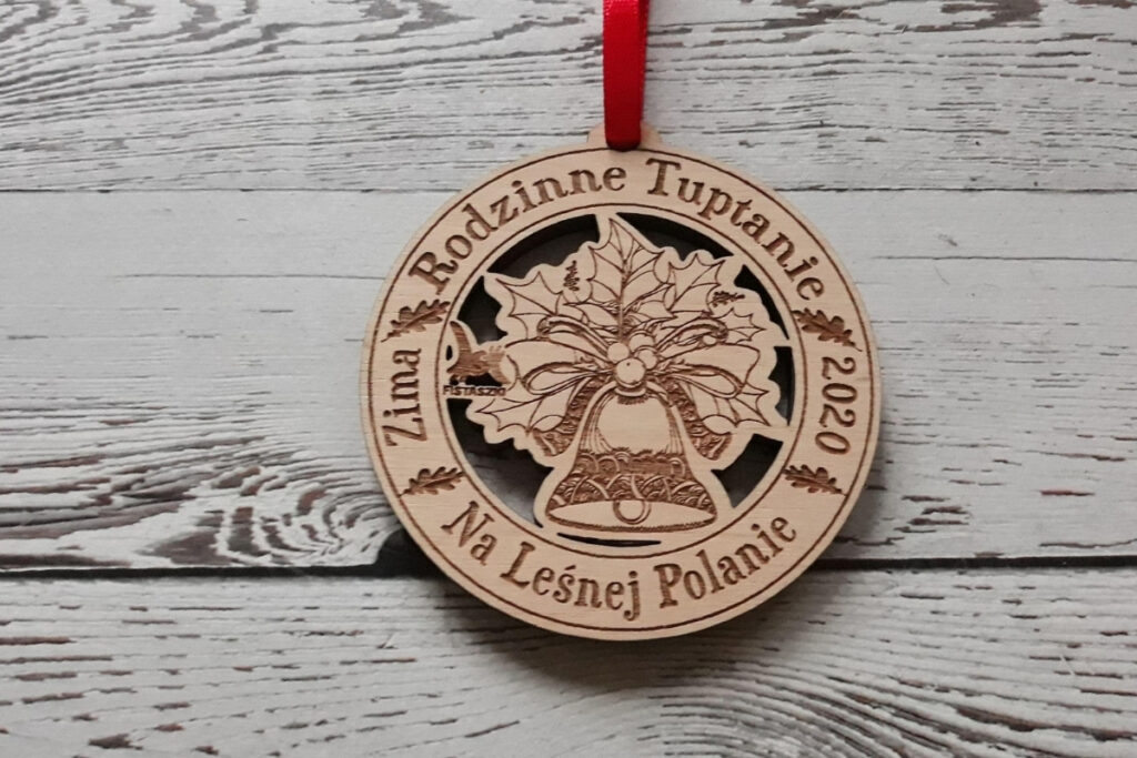 Okrągły drewniany medal na bieg „Rodzinne Tuptanie”, z precyzyjnie wyciętym ażurowym dzwonkiem – symbolem Wielkanocy – oraz grawerem liści i napisów , wykonany z pełnej płyty jasnej brzozy bez osmaleń, bez sęków, grubość medalu 6mm. Medal na czerwonej wstążce.