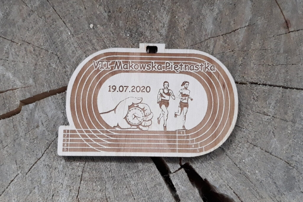 Drewniany medal na bieg „Makowska Piętnastka”, z grawerem imitującym tory bieżni lekkoatletycznej, sylwetką biegaczy oraz ręką trzymającą stoper - mierzącą czas, wykonany z czystej brzozy bez osmaleń.
