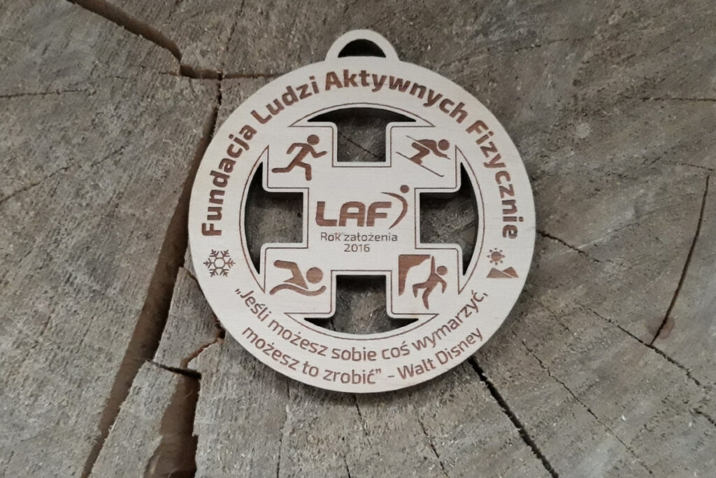Drewniany okrągły medal z ażurem w środku. Piktogramy narciarza, biegacza, pływaka i alpinisty. W środku także logo Fundacji.