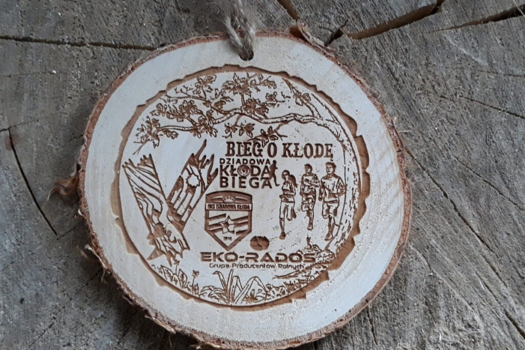 Naturalny drewniany medal na „V Bieg o Kłodę”, wykonany na autentycznym plastrze brzozy z korą, z grawerem drzewa, biegaczy oraz logotypem, zachowujący jasny odcień drewna bez osmaleń.