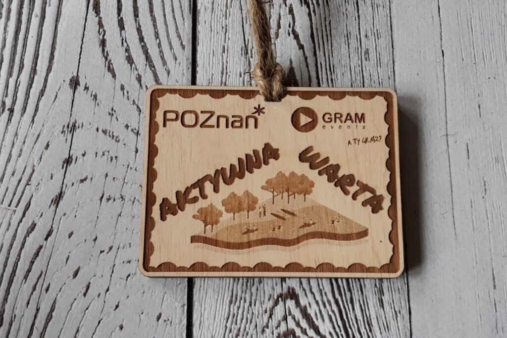 Prostokątny drewniany medal projektu „Aktywna Warta”, z grawerem wybrzeża na którym są kajaki, promującym lokalną aktywność, wykonany z czystej brzozy bez osmaleń.