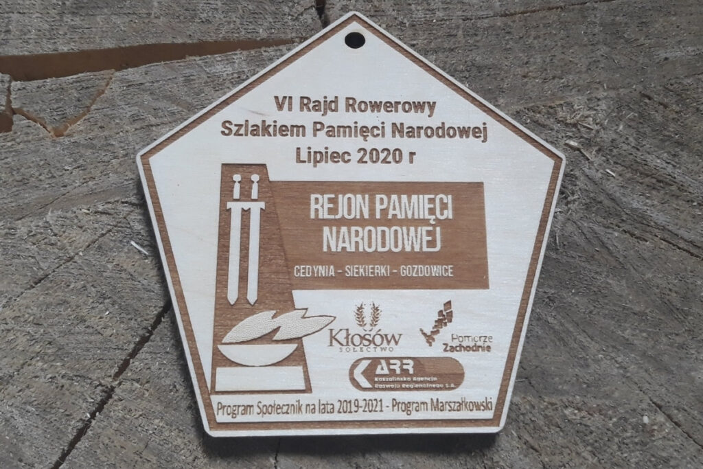 5 - kątny drewniany medal upamiętniający „Rajd Rowerowy Szlakiem Pamięci Narodowej”, z grawerem historycznych symboli, wykonany z jasnej brzozy o nieskazitelnie czystej powierzchni bez osmaleń. Na medalu logotypy.