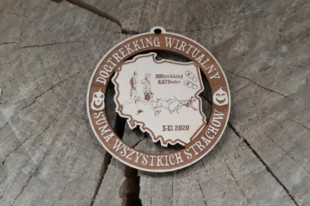 Okrągły drewniany medal na „Dogtrekking Wirtualny”, z precyzyjnie wyciętym ażurowym konturem Polski, grawerem sylwetek 2 biegaczy z psem, wykonany z jasnej płyty brzozy bez osmaleń.