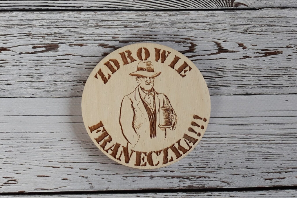 Okrągła drewniana podstawka pod piwo z grawerem osoby pijącej piwo, oraz personalizowanym napisem „Zdrowie Franeczka”, wykonana z jasnej płyty brzozy o idealnie czystej powierzchni bez śladów osmaleń.