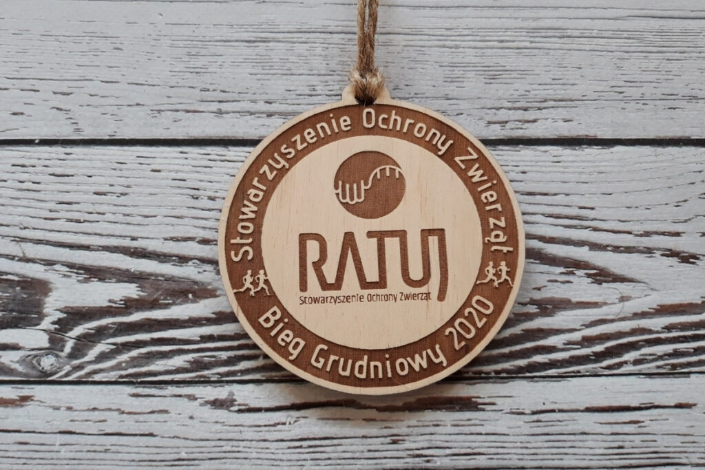 Okrągły drewniany medal dla Stowarzyszenia Ochrony Zwierząt „Ratuj”, z precyzyjnie wygrawerowanym logo stowarzyszenia w części centralnej, wykonany z jasnej brzozy o idealnie czystej powierzchni bez śladów osmaleń.