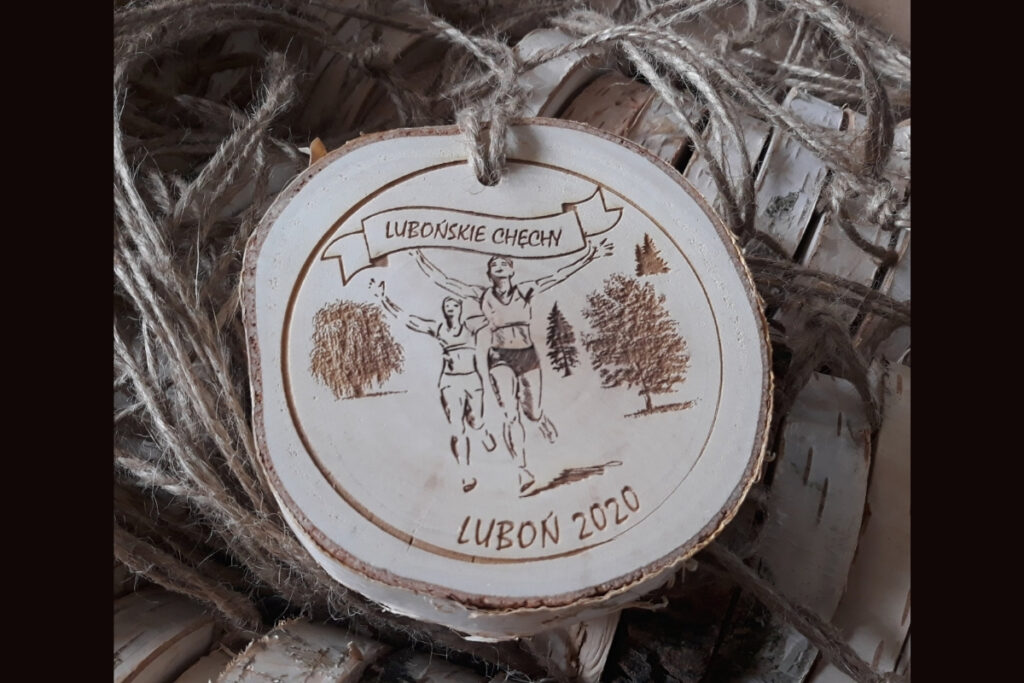 Drewniany medal na plastrze brzozy z napisem Lubońskie Chęchy. pomiędzy drzewami biegnie 2 biegaczy, w rękach trzymają baner z tym napisem. Medal na sznurku jutowym.