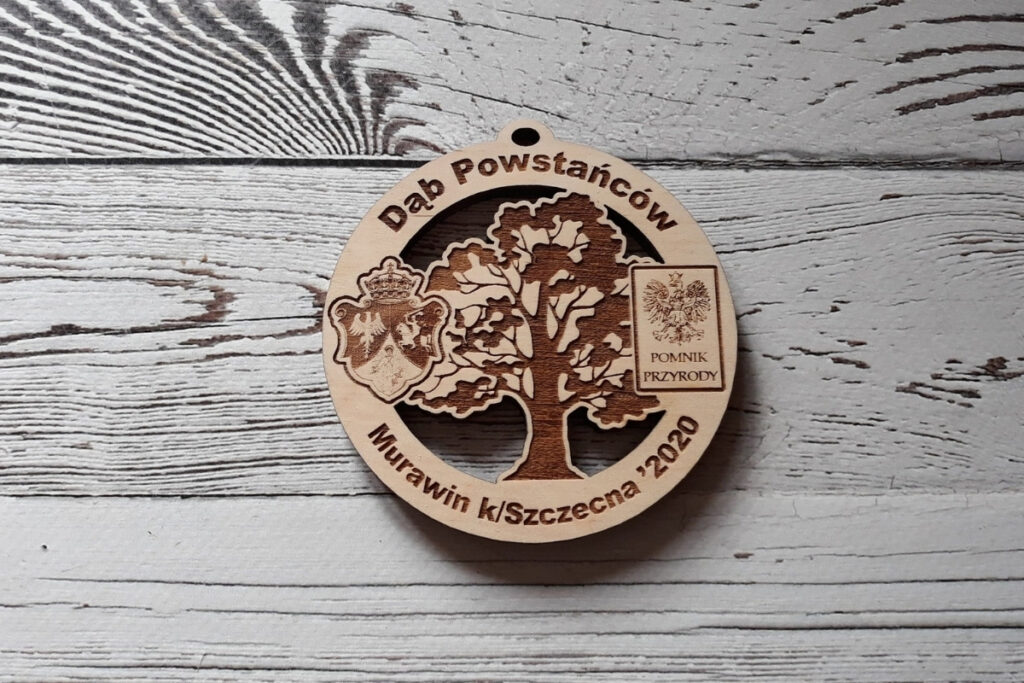 Okrągły drewniany medal z misternie grawerowanym i ażurowo wyciętym wizerunkiem „Dębu Powstańców”, z dbałością o detale korony i pnia, wykonany z czystej brzozy bez osmaleń. Na medalu są także 2 logotypy.