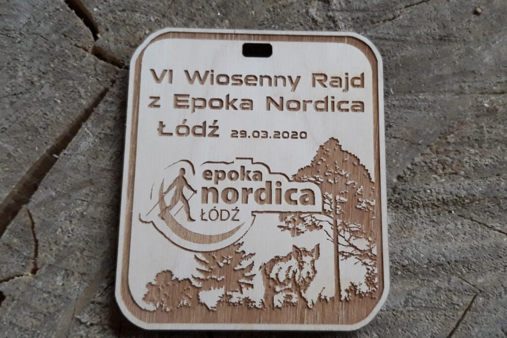 Prostokątny medal z logiem Epoka Nordica, dzikiem oraz drzewami.