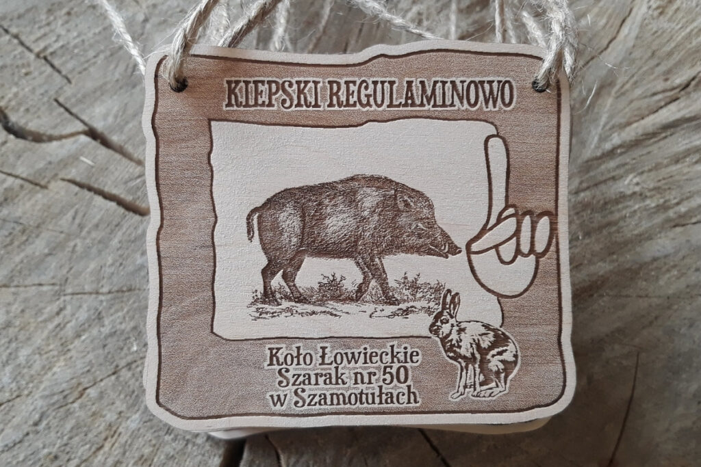 Kwadratowy, prestiżowy medal myśliwski o grubości 6 mm, wykonany z litej sklejki brzozowej, z grawerem dzika oraz dłoni z palcem wskazującym dla Kiepskiego Regulaminowo.
