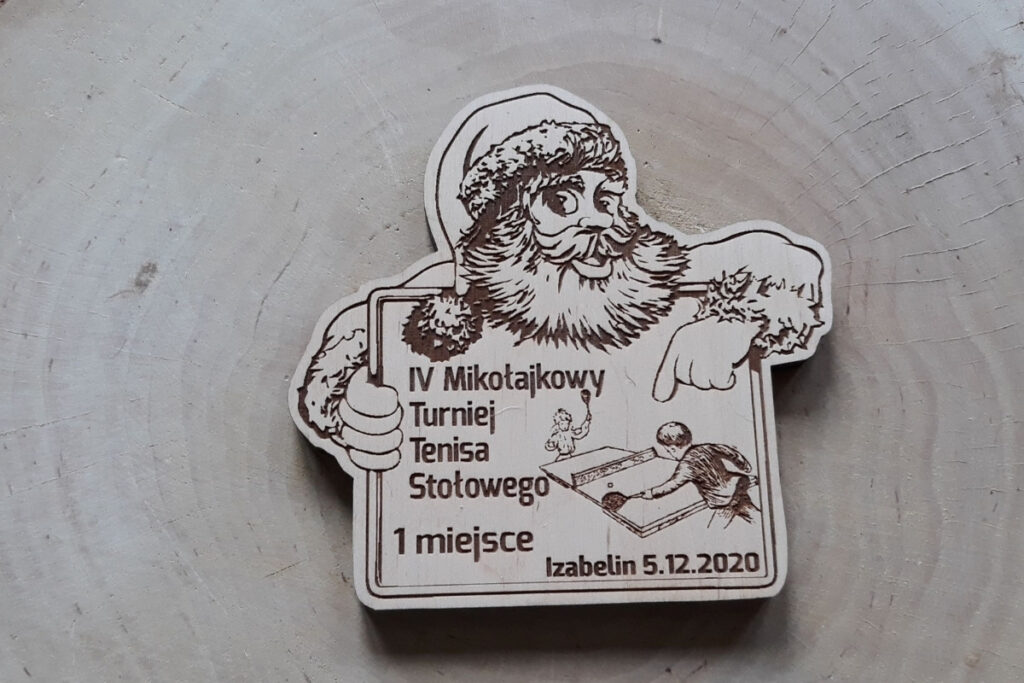 Drewniany medal na „Mikołajkowy Turniej Tenisa Stołowego”, w kształcie mikołaja trzymającego tablicę, z grawerem 2 osób grających w ping-ponga oraz świątecznej czapki Mikołaja, wykonany z czystej brzozy bez osmaleń.