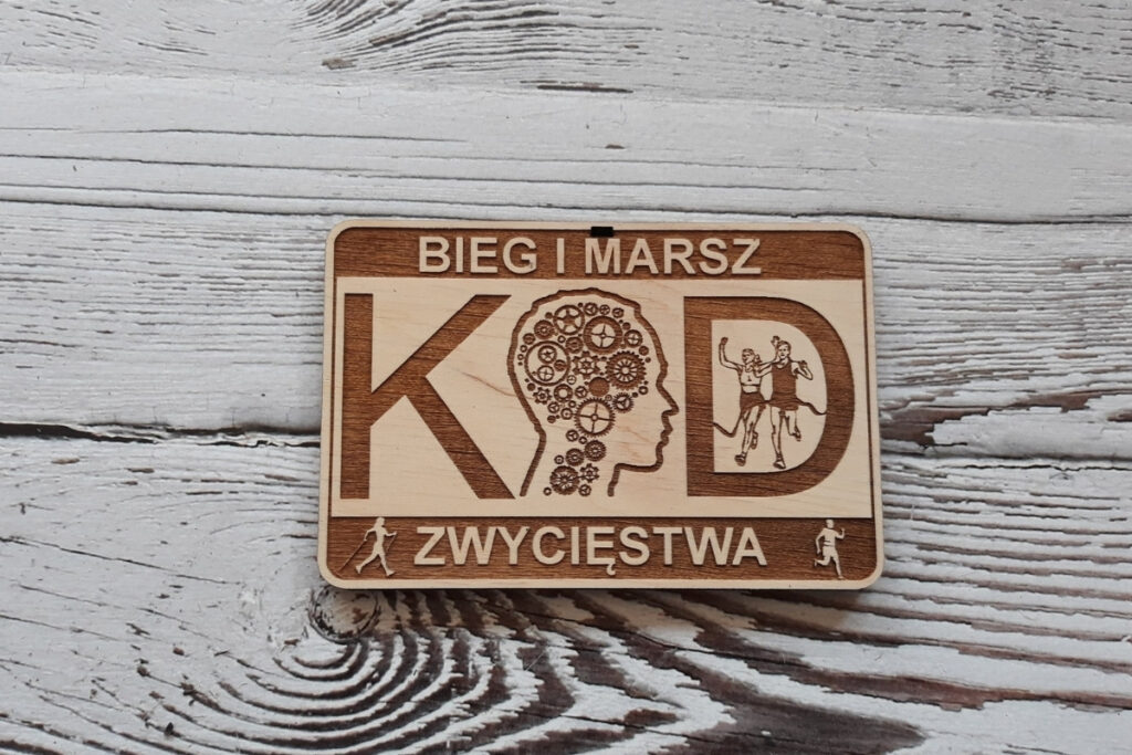 Prostokątny medal z napisami Kod Zwycięstwa Bieg i Marsz. w napisie KOD zamiast litery O jest głowa z trybikami.