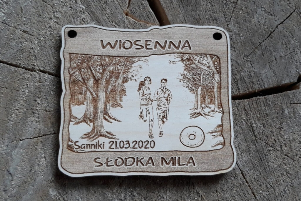 Prostokątny drewniany medal na bieg Wiosenna Słodka Mila z grawerem drzew po bokach, biegaczami w środku i apetycznego pączka na przodzie w leśnej oprawie.