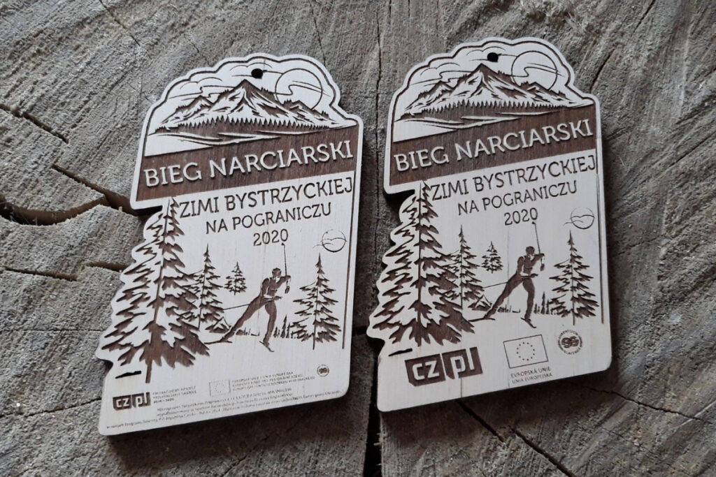 Drewniany medal na Bieg Narciarski Ziemi Bystrzyckiej z grawerem narciarza na tle górskich świerków, u góry nad napisem pasmo górskich  szczytów.