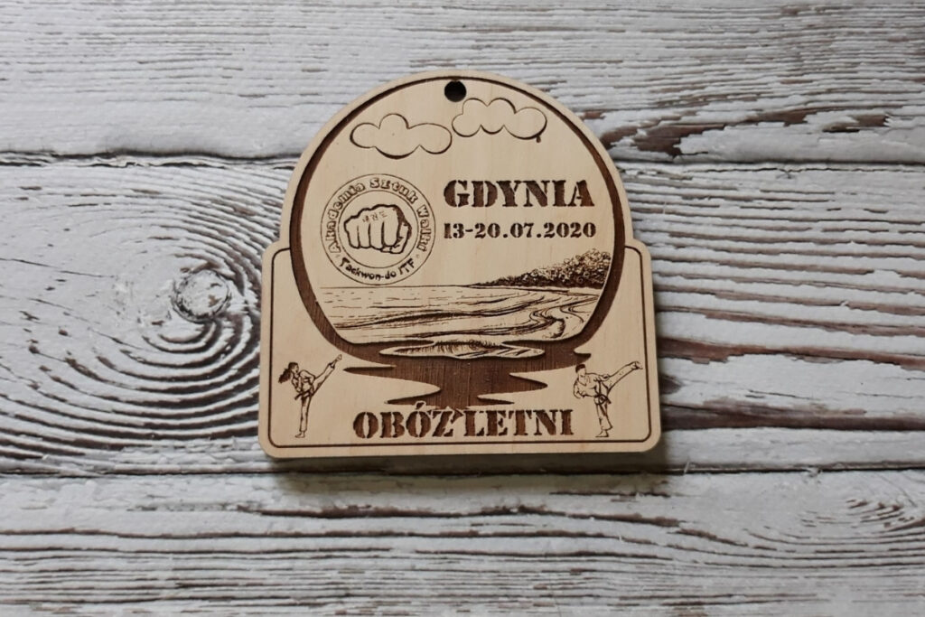 Półokrągły drewniany medal pamiątkowy z obozu letniego w Gdyni, z grawerem fal i brzegu,  wykonany z jasnej brzozy bez śladów osmaleń.
