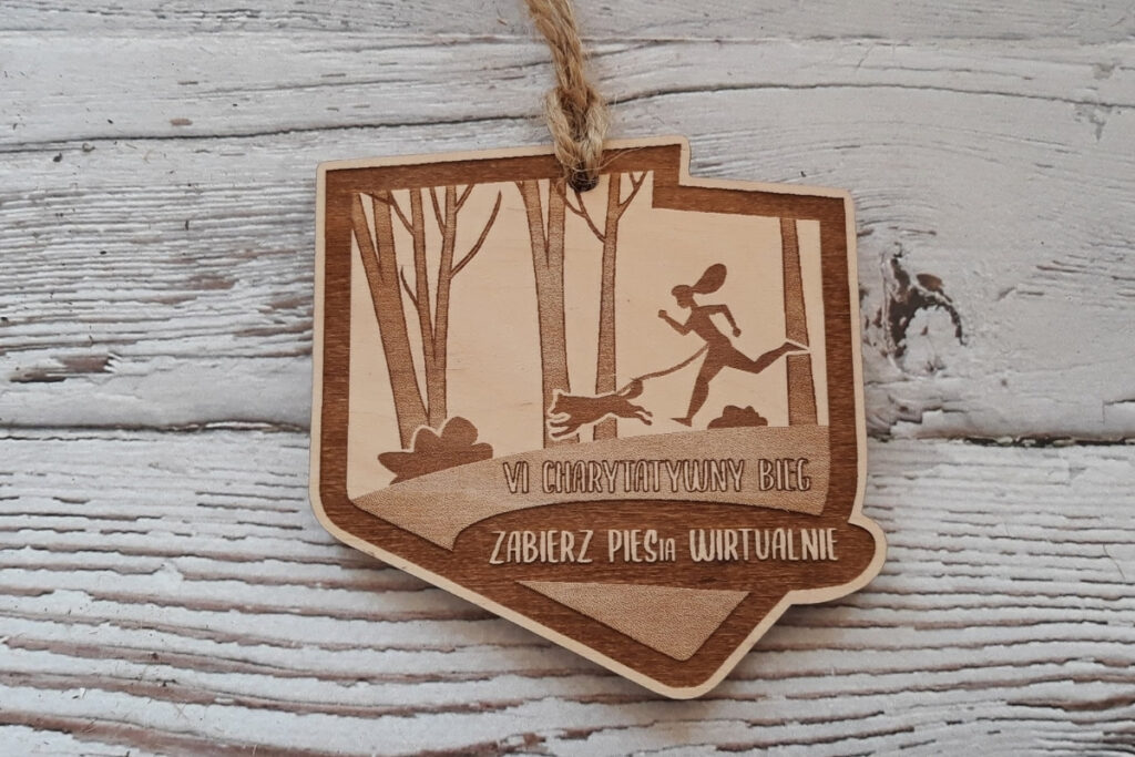 Drewniany medal charytatywny w kształcie konturów Polski z grawerem psa i napisem „Zabierz piesia wirtualnie”.