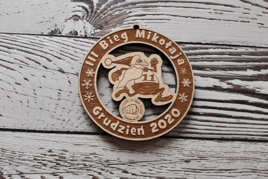 Okrągły ażurowy drewniany medal na Bieg Mikołaja z wyciętym motywem biegnącego mikołaja w czapce mikołajkowej i radosnym grawerem. Na medalu widnieje także logotyp. Napis to III Bieg Mikołaja grudzień 2020.