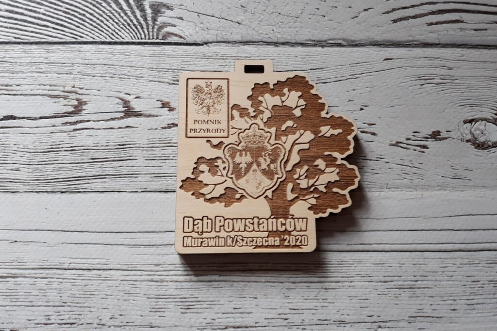 Historyczny drewniany medal z grawerem rozłożystego dębu powstańców i logotypami. Dąb jest Pomnikiem Przyrody.