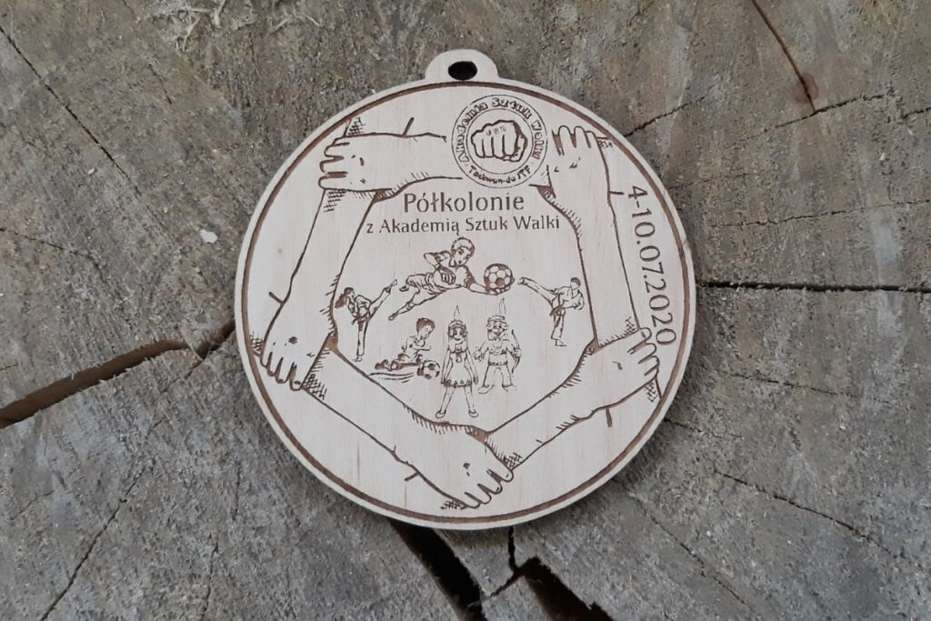 Okrągły drewniany medal na letnie półkolonie z inspirującym grawerem ramki w postaci trzymających się wzajemnie dłoni. Na medalu napis półkolonie i różne dyscypliny sportu, oraz logotyp.