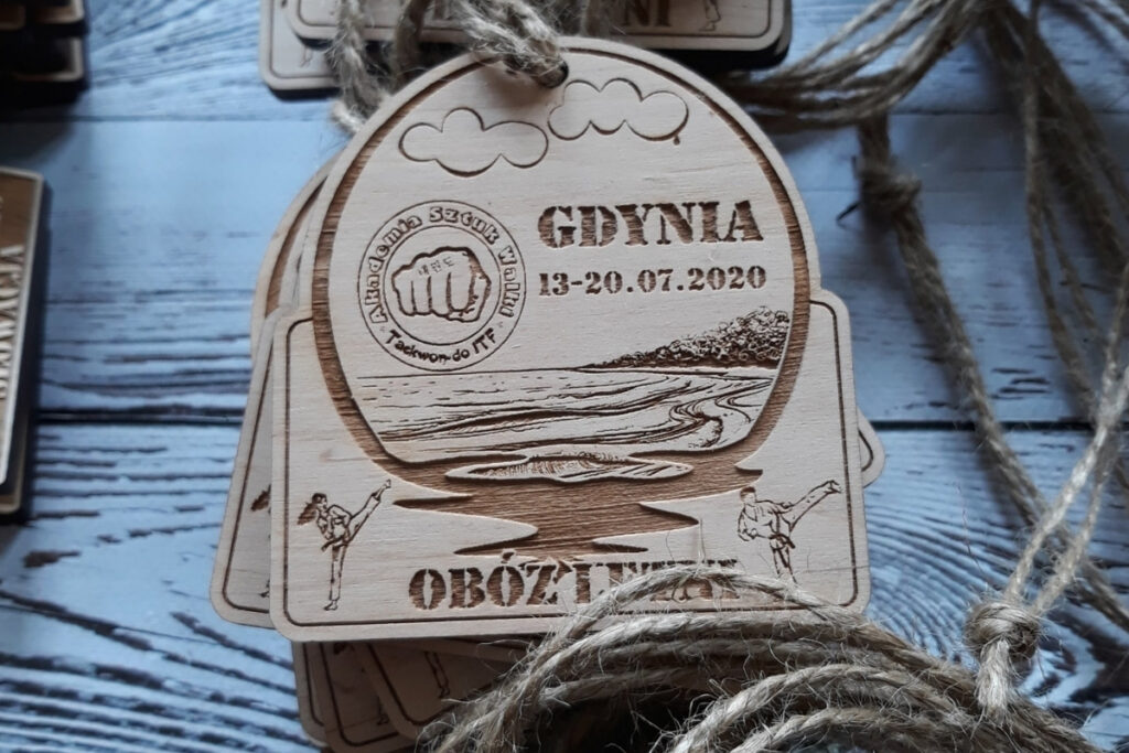 Półokrągły drewniany medal pamiątkowy z obozu letniego w Gdyni, z grawerem fal i brzegu,  wykonany z jasnej brzozy bez śladów osmaleń.