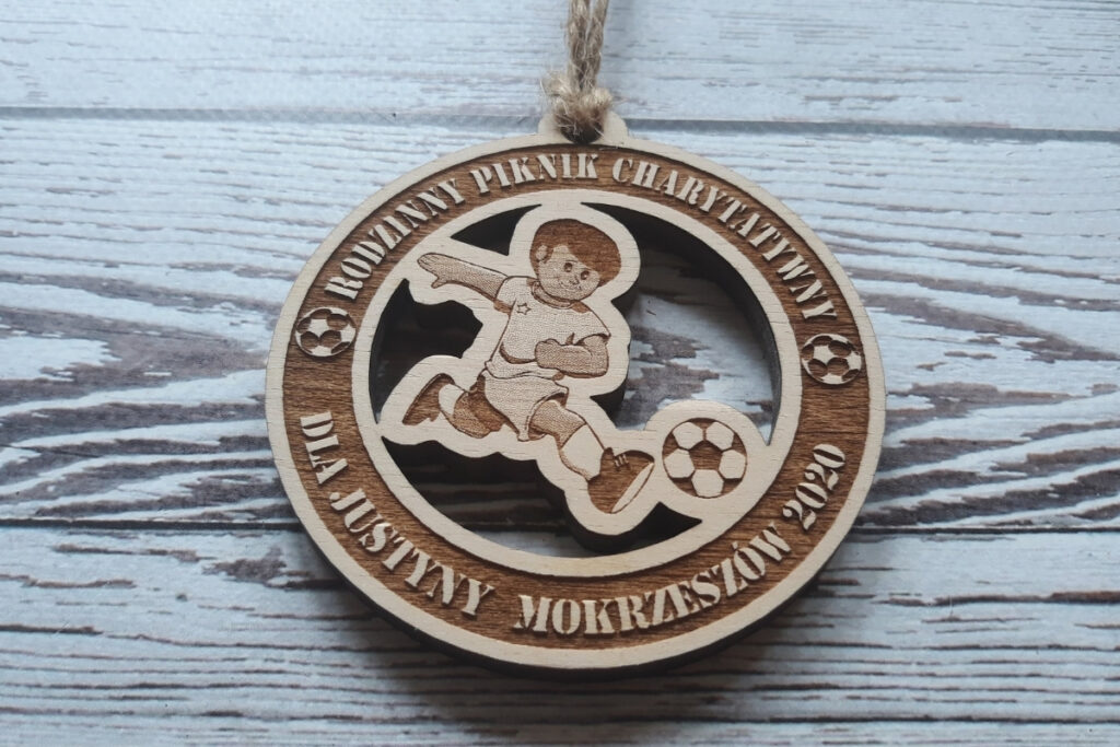 Okrągły drewniany medal na Rodzinny Piknik Charytatywny z ażurowym wycięciem postaci młodego piłkarza i grawerem pamiątkowym.