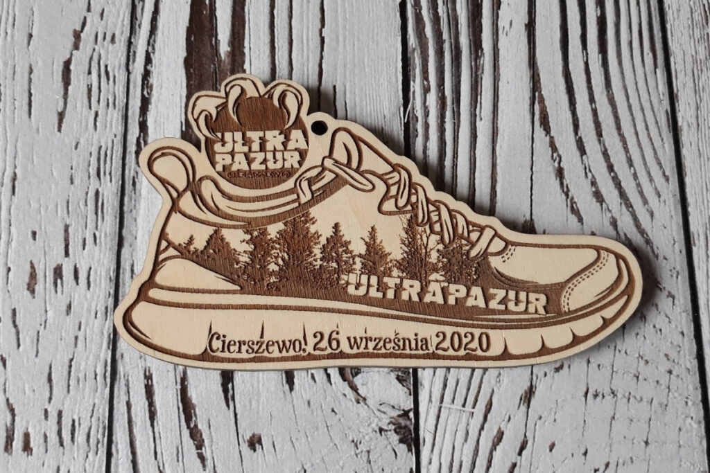 Oryginalny drewniany medal wycięty w kształcie buta sportowego z precyzyjnym grawerem sznurowadeł i detali biegowych. Na bucie wygrawerowany las, u góry logotyp.