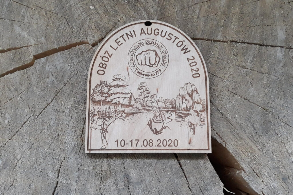 Drewniany medal na obóz letni w Augustowie z grawerem kajaka na jeziorze i pamiątkowym napisem.
