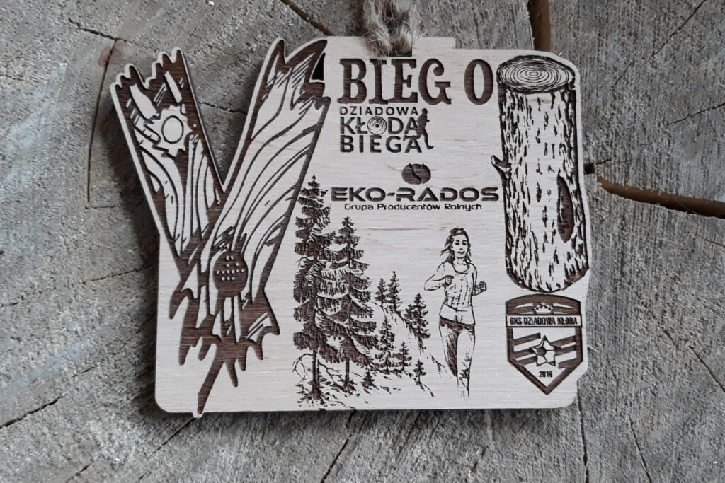 Drewniany medal na jubileuszowy V Bieg o Kłodę z dużą rzymską cyfrą V oraz grawerem kłody drewna, biegacz, lasku i logotypu.