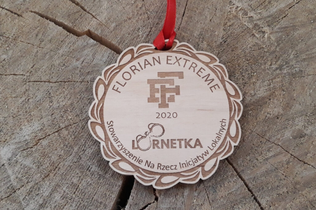 Okrągły drewniany medal na wstążce z elegancką, grawerowaną ramką i centralnie umieszczonymi logotypami organizatorów.